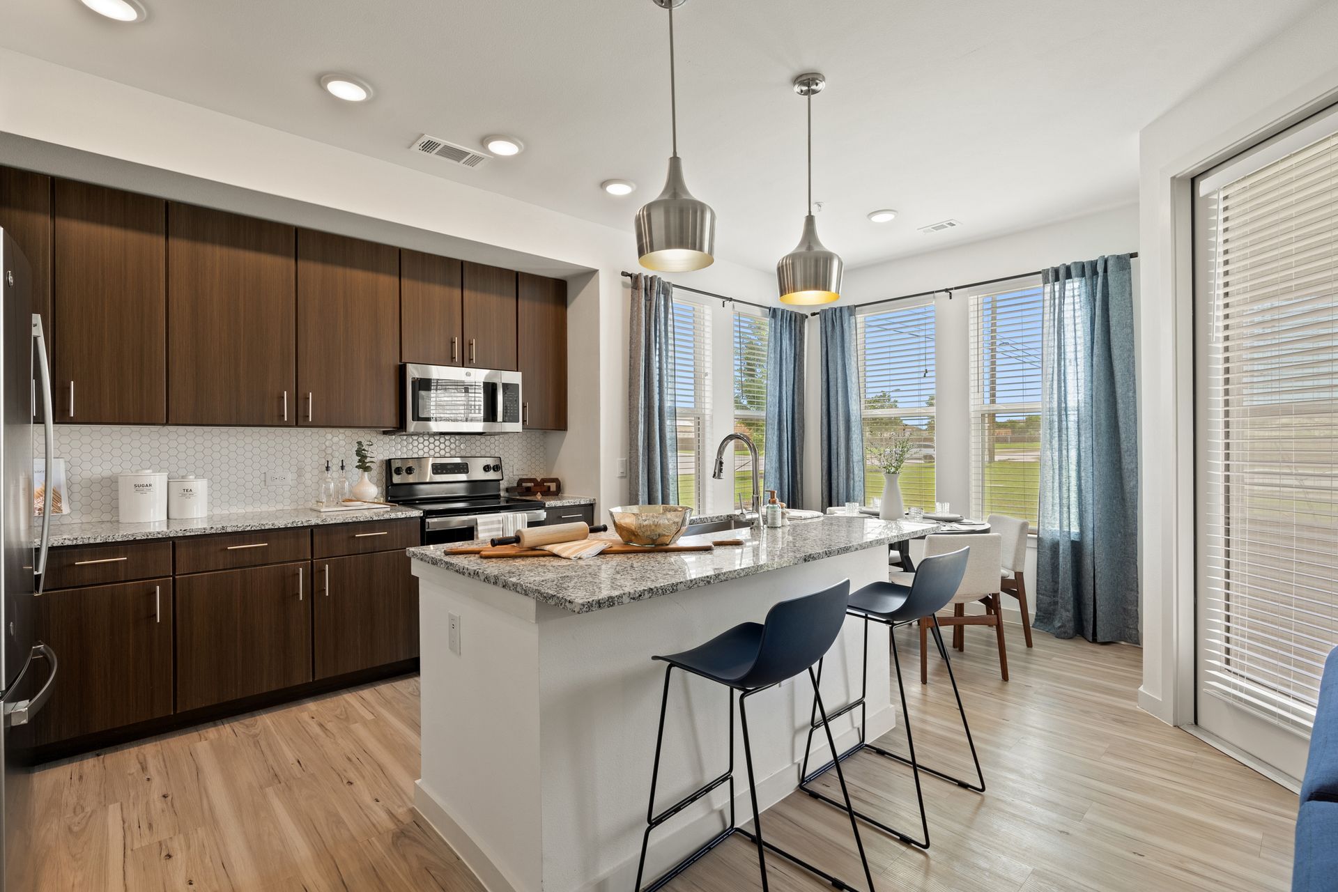 Modern kitchen with dark wood cabinets, granite island, stainless appliances, and a sunny dining area by windows.