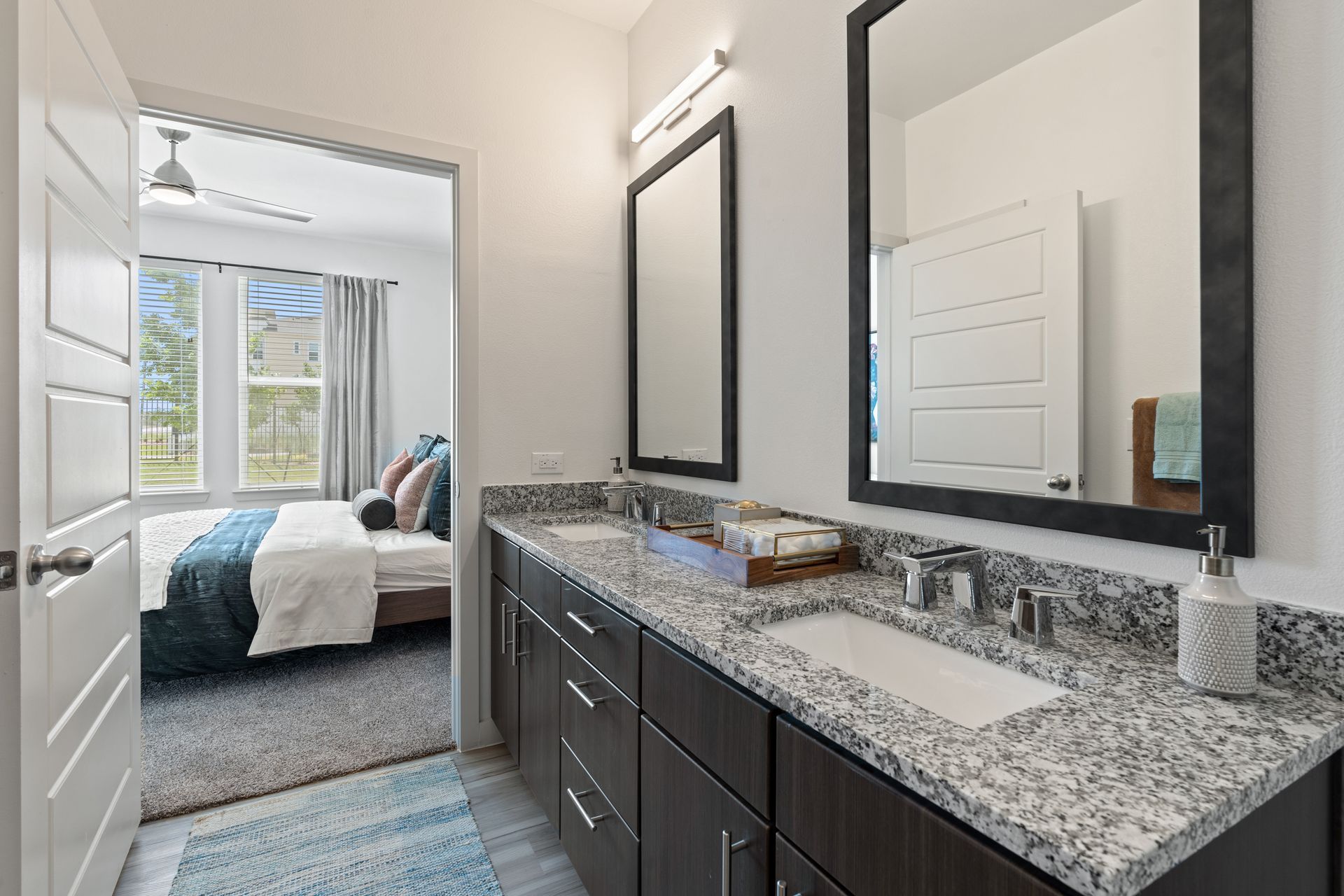 Bathroom vanity with granite countertop, dual sinks, and two framed mirrors; doorway to a bedroom.