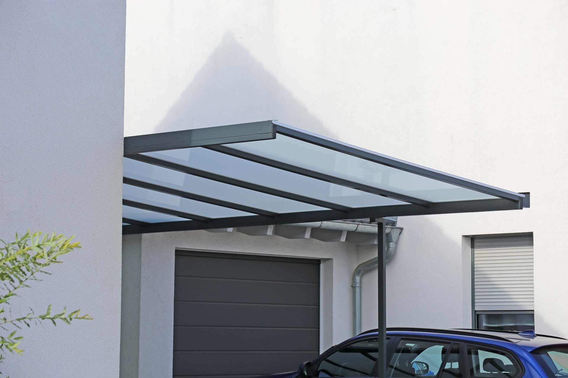 A blue car is parked under a canopy in front of a garage.