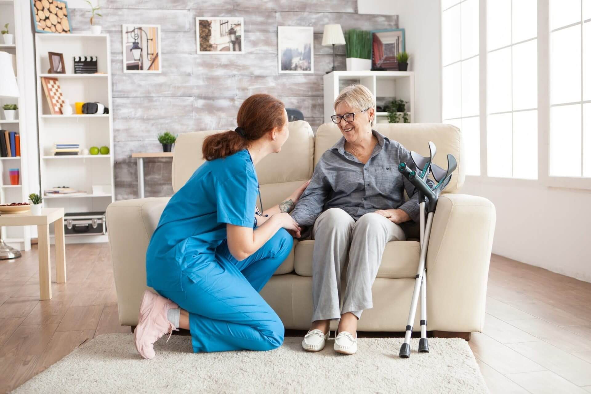 A nurse is kneeling down next to an elderly woman sitting on a couch.