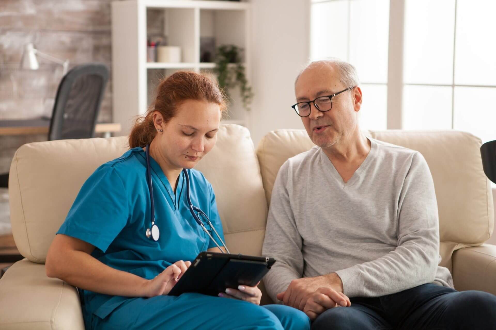 A nurse is sitting on a couch with an elderly man and looking at a tablet.