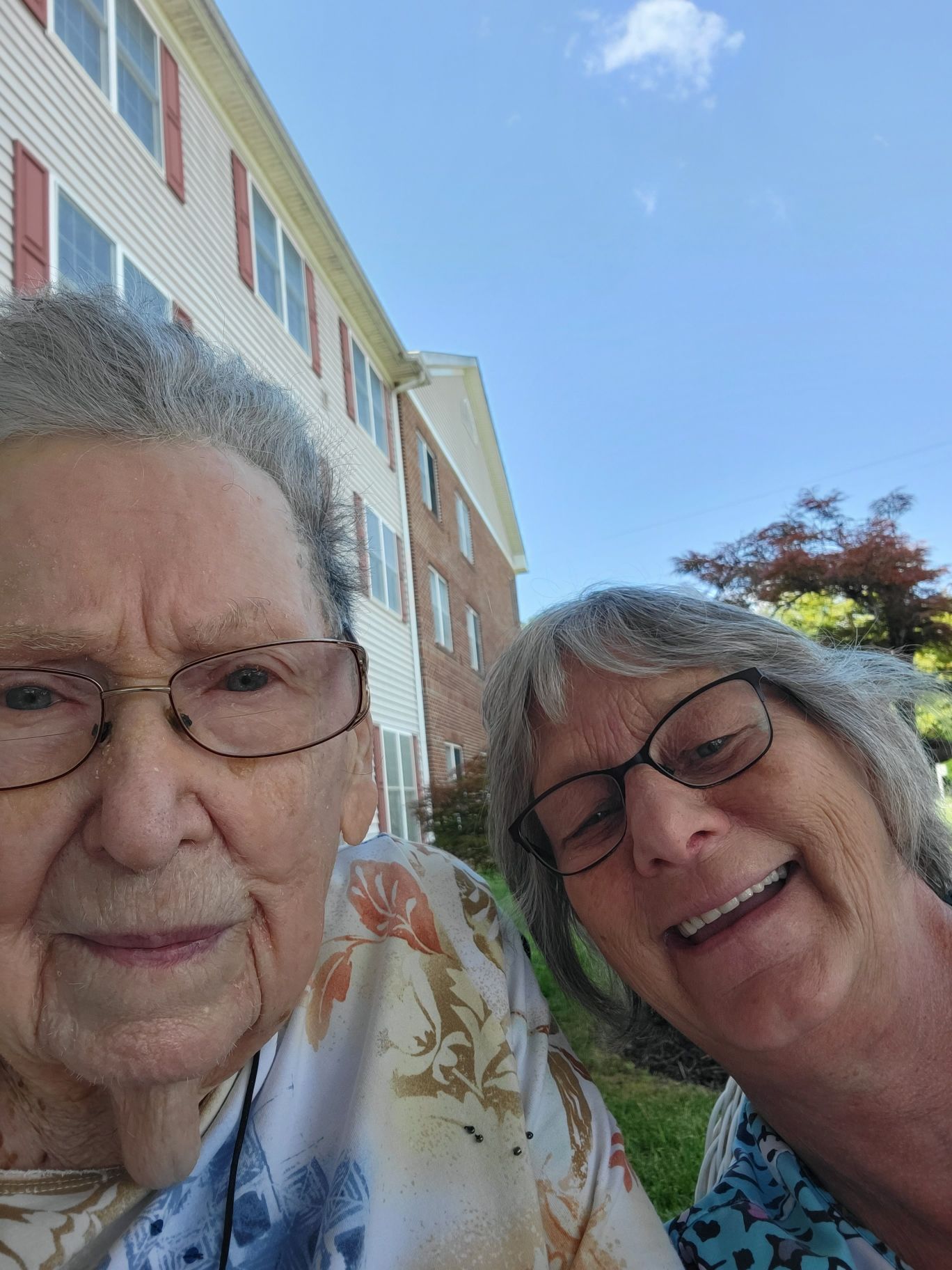 Two older women are taking a selfie in front of a building.