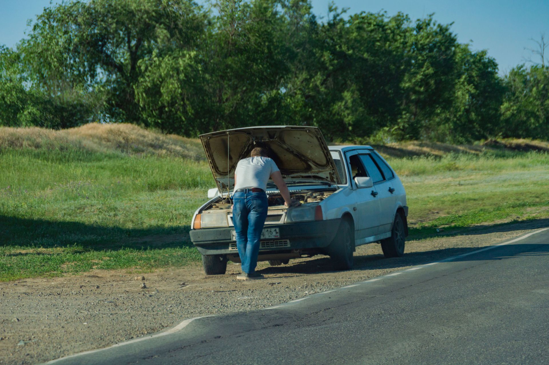 Person with open hood of a blue car parked on a roadside to overheating engine | Auto Care Unlimited