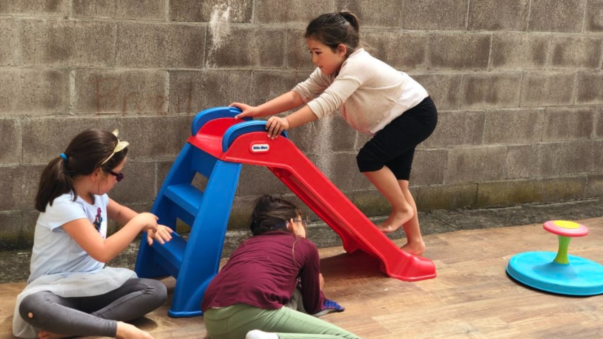 Children playing on a red-and-blue slide in a courtyard, with a toy spinner nearby