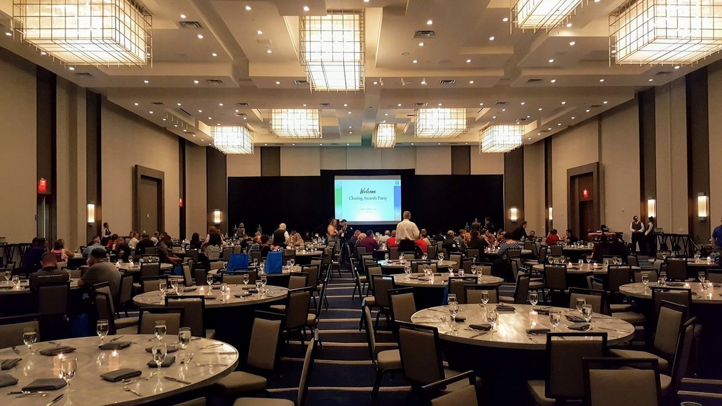 Large conference room with round tables and people seated, watching a screen.