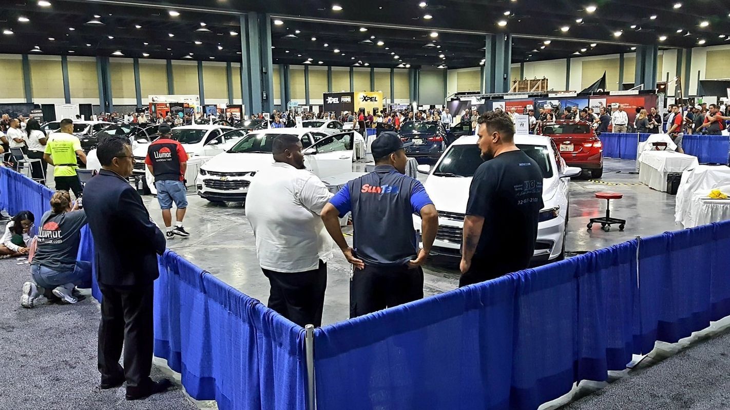Car show inside a large hall with attendees and vehicles. Blue barriers, people standing and viewing.