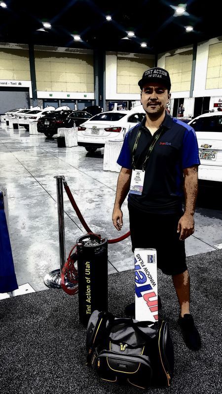 Man standing with gear at a car show; black uniform, black bag, soda canister, cars in background.