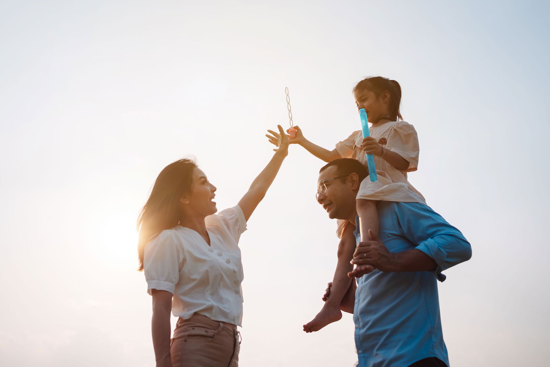 Family playing outdoors. Daughter on father's shoulders blowing bubbles, mother reaching up. Backlit by the sun.
