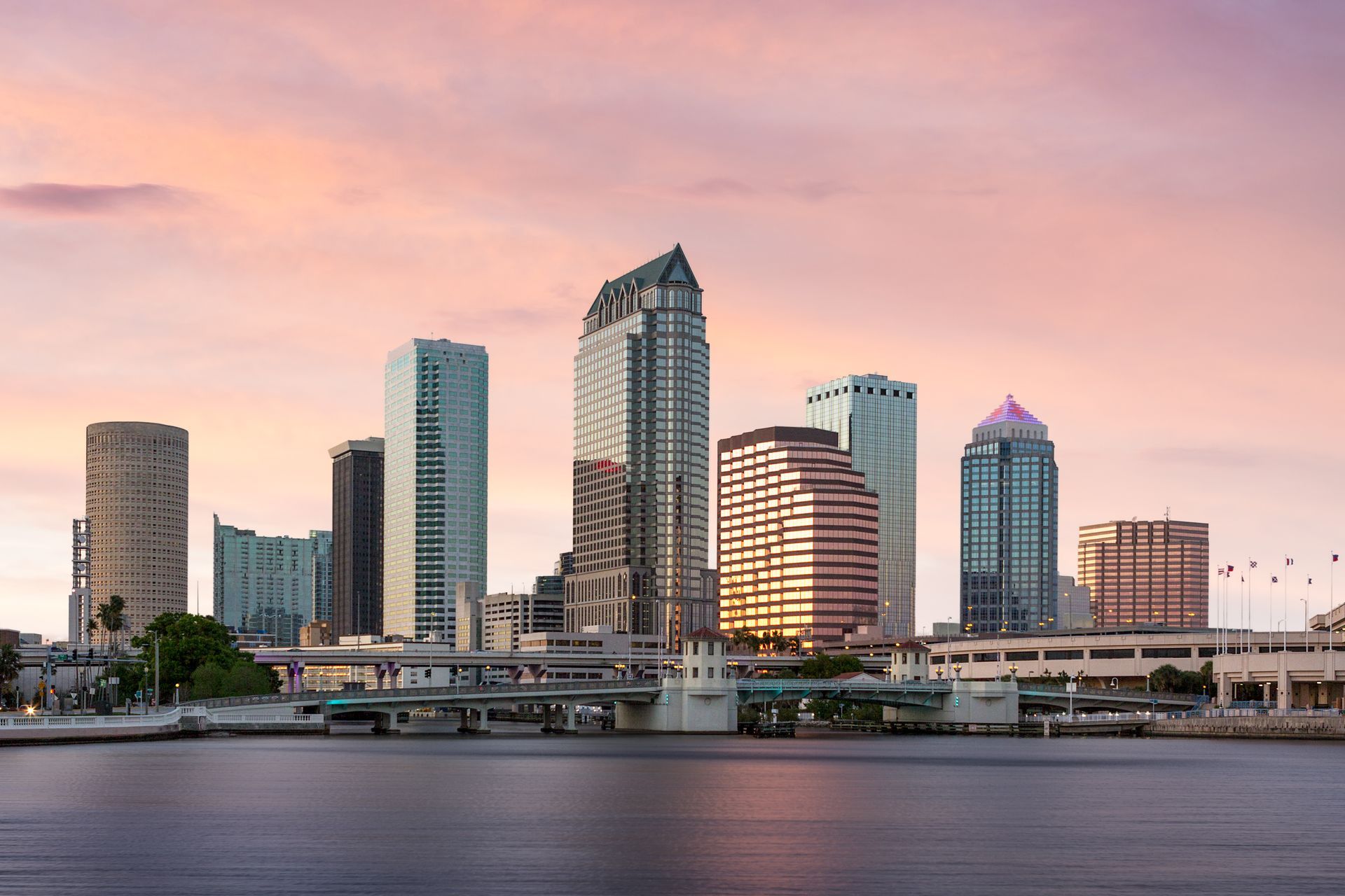 Tampa, Florida skyline at sunset, with high-rise buildings silhouetted against a pink and orange sky, reflecting in the water.