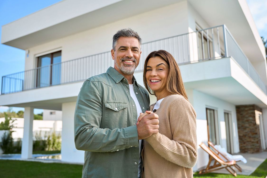 Smiling couple stands in front of a modern white house, holding hands and looking at the camera. Green lawn, blue sky.