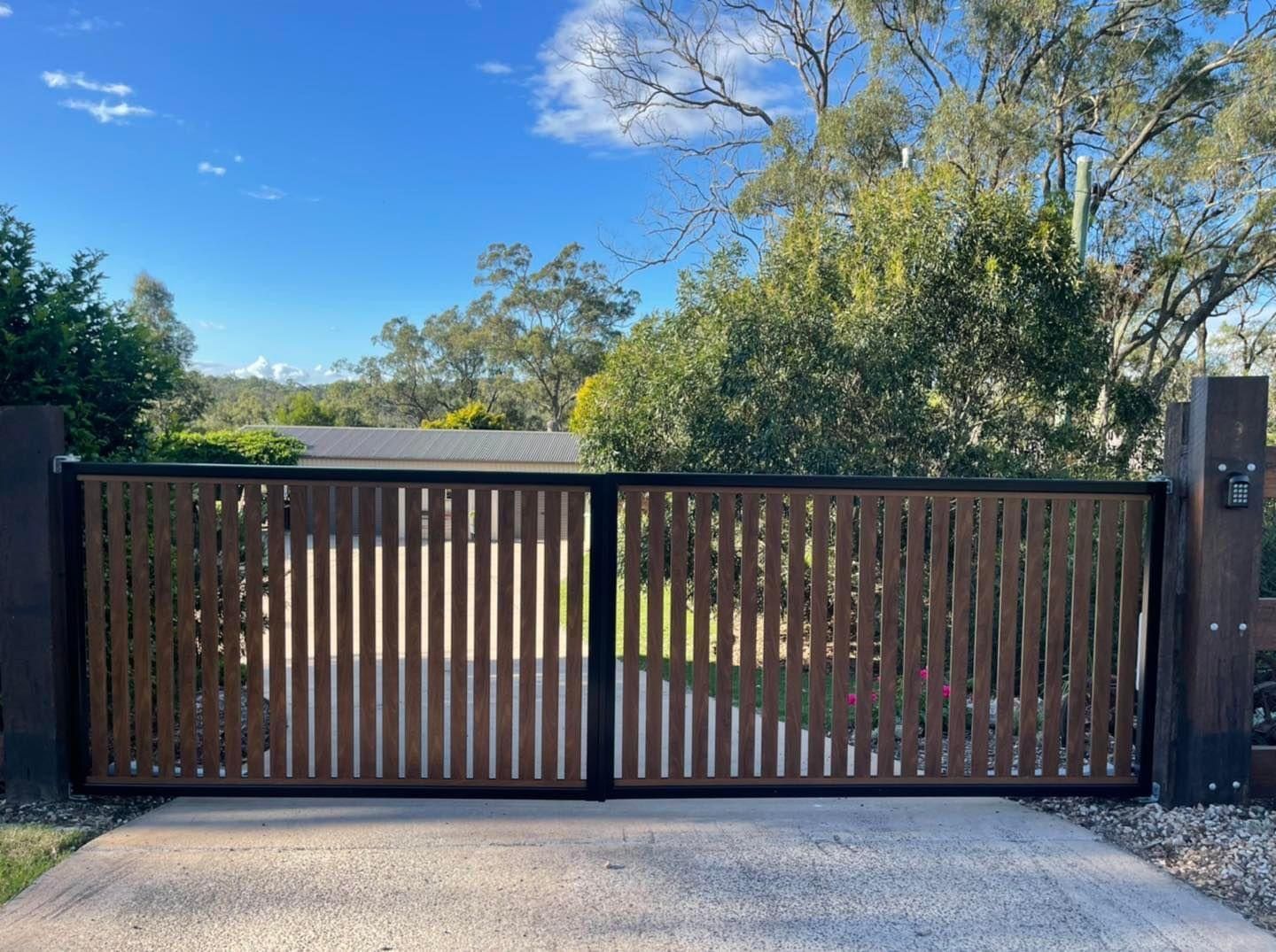 A wooden gate is open to a driveway with trees in the background.