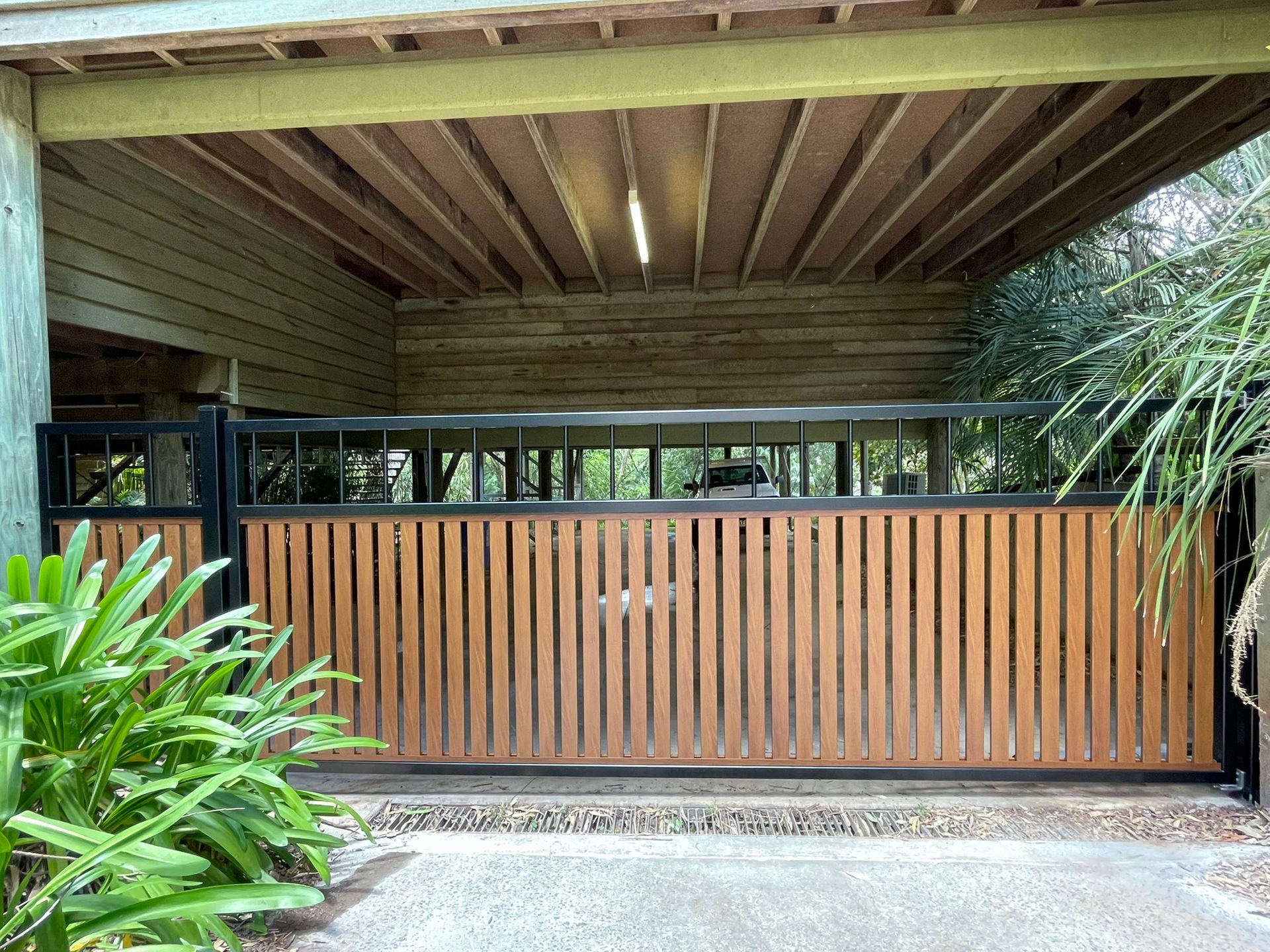 A wooden gate is sitting under a wooden roof next to a driveway.