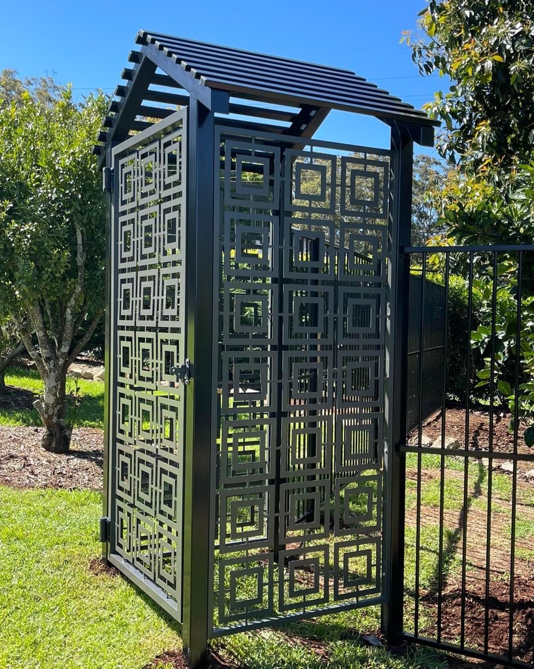 A black gate with a laser cut geometric pattern on it is sitting in the grass next to a fence.