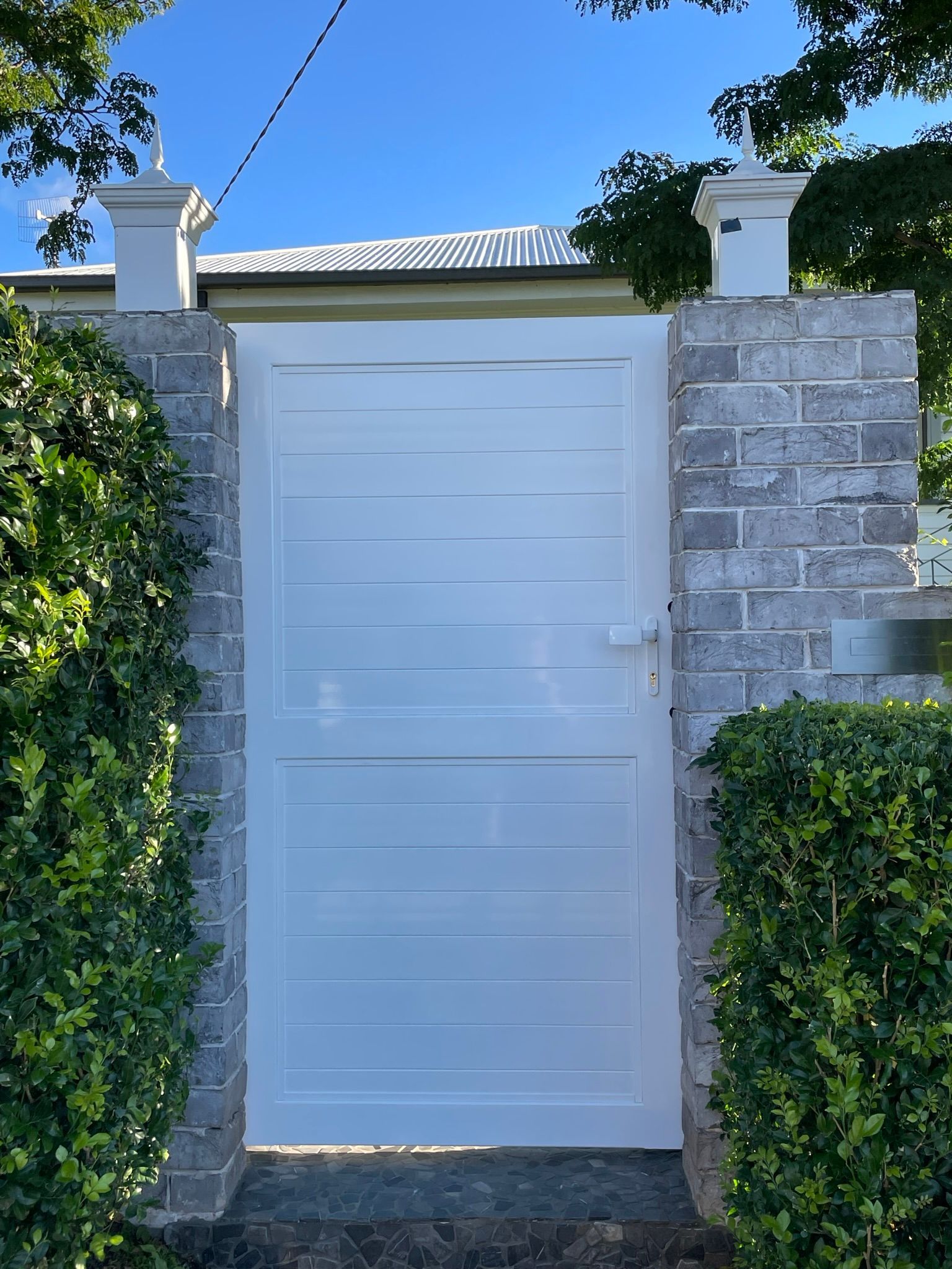 A white gate is surrounded by a brick wall and bushes.