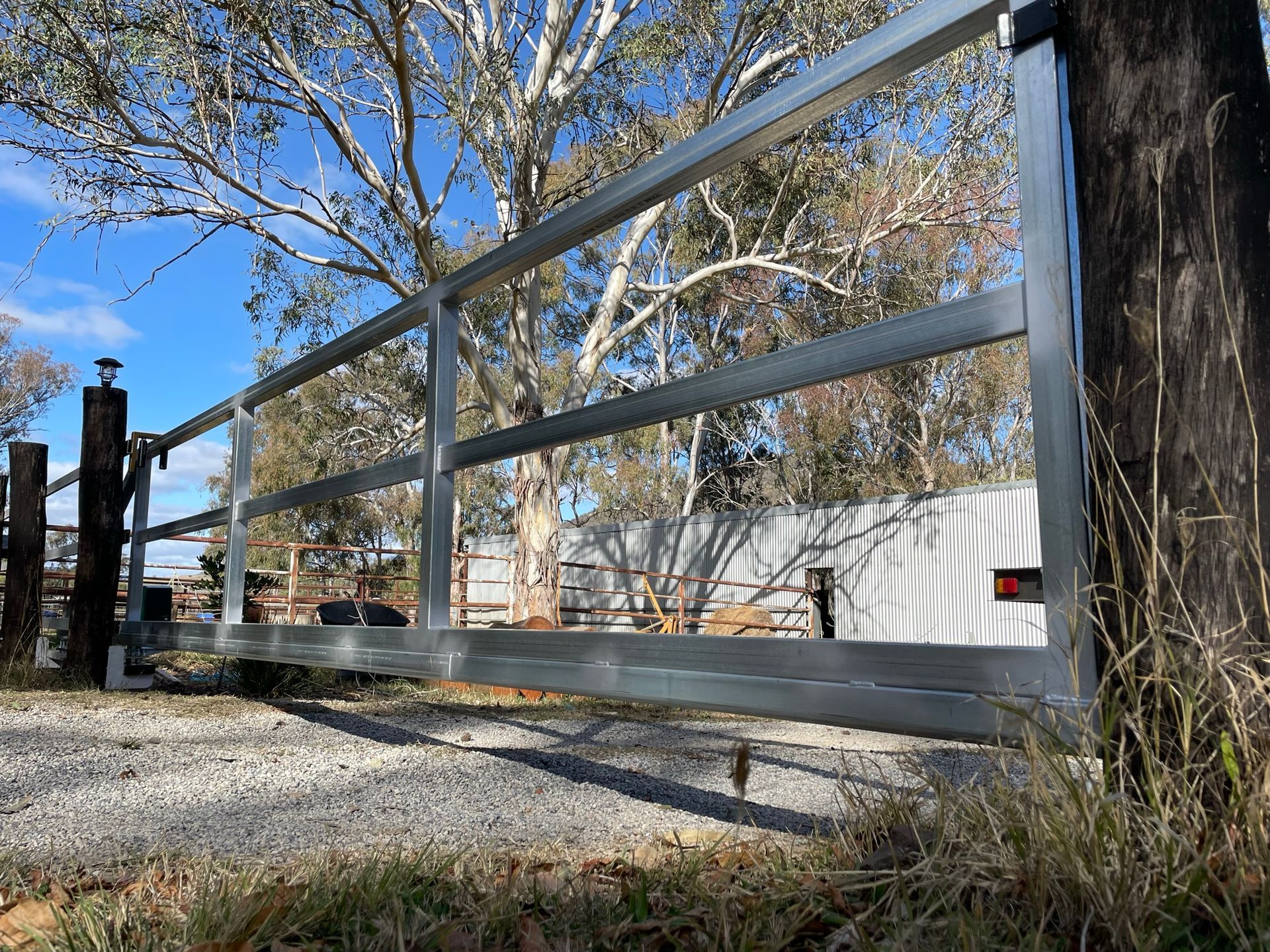 A metal gate is sitting next to a tree on a gravel road.