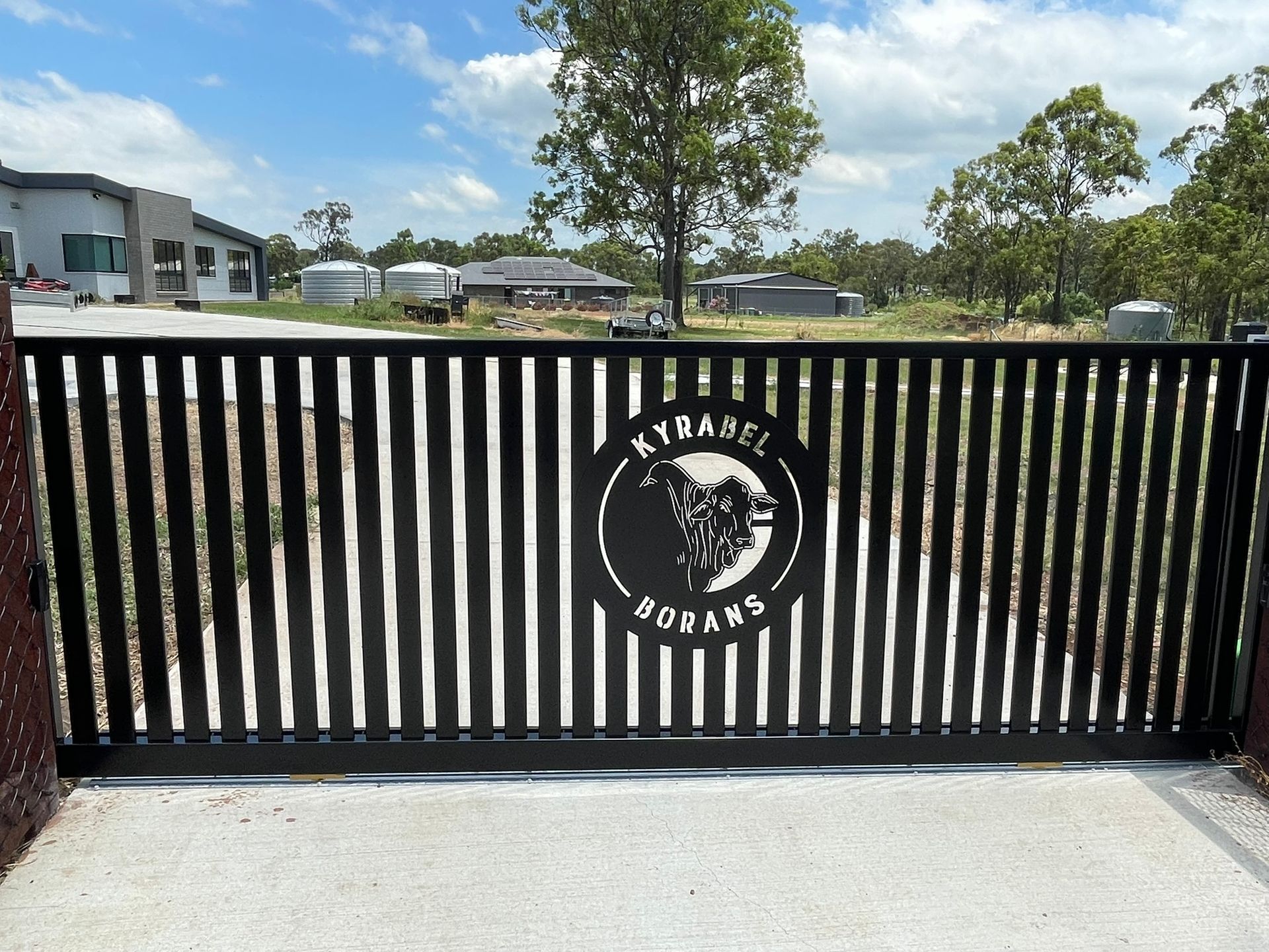 A metal gate is sitting next to a tree on a gravel road.