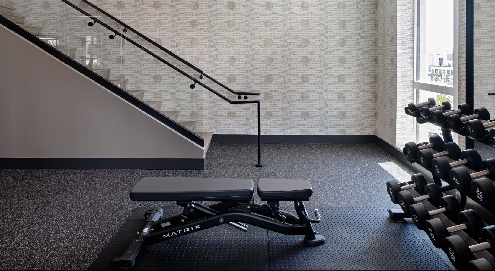 Indoor apartment gym with a weight bench, dumbbells, and a staircase beside a large window at Sofia, offers Santa Clara apartments for rent.