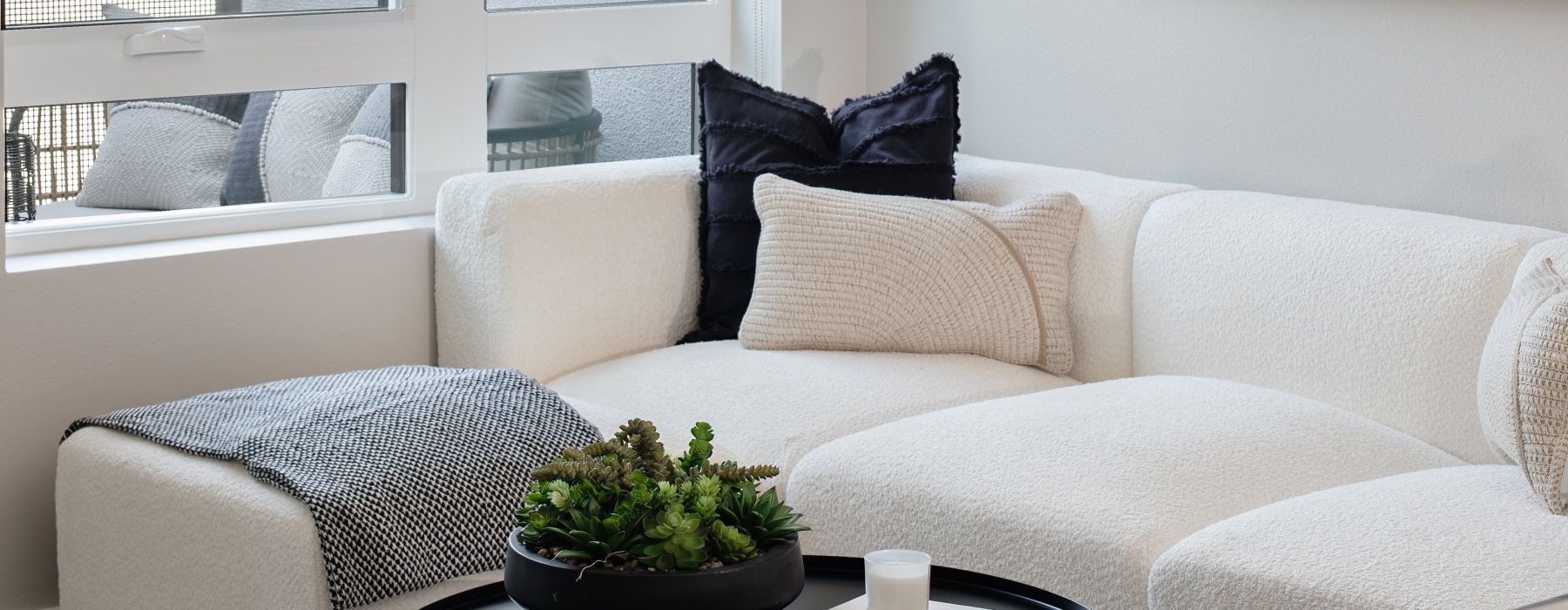 White sectional sofa with beige cushions near a window, a plant and candle on a black tray at Sofia, offers luxury apartment living in Santa Clara, CA.