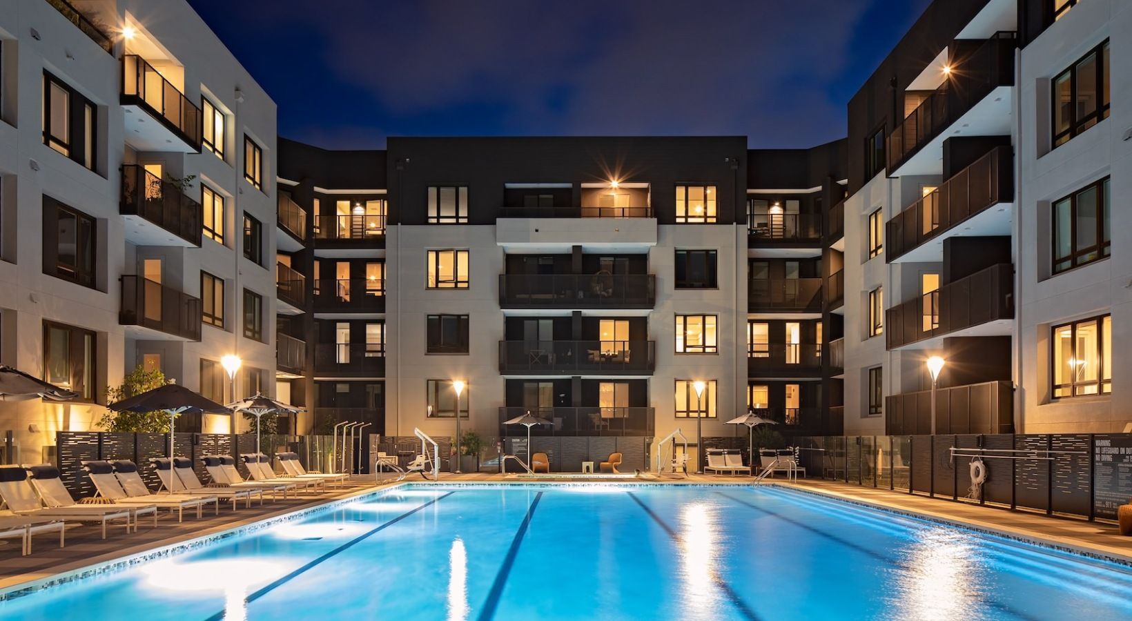 Nighttime view of a modern apartment complex with a lit swimming pool and balconies at Sofia, offers Santa Clara apartments for rent in CA.