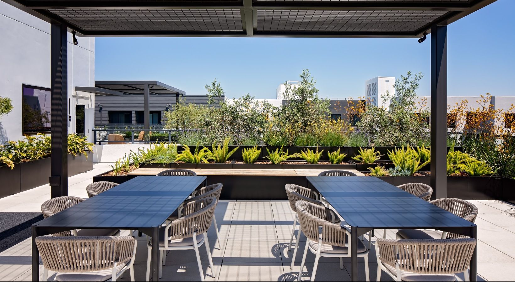 Outdoor rooftop courtyard with two long dining tables, woven chairs, and lush planters at Sofia, offers apartments in Santa Clara, CA.