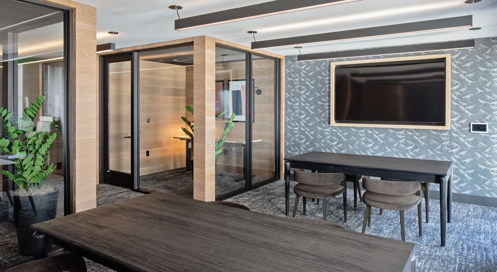 Modern community lounge with glass partitions, tables, plants, and a wall-mounted TV at Sofia, offers Santa Clara apartments for rent in CA.