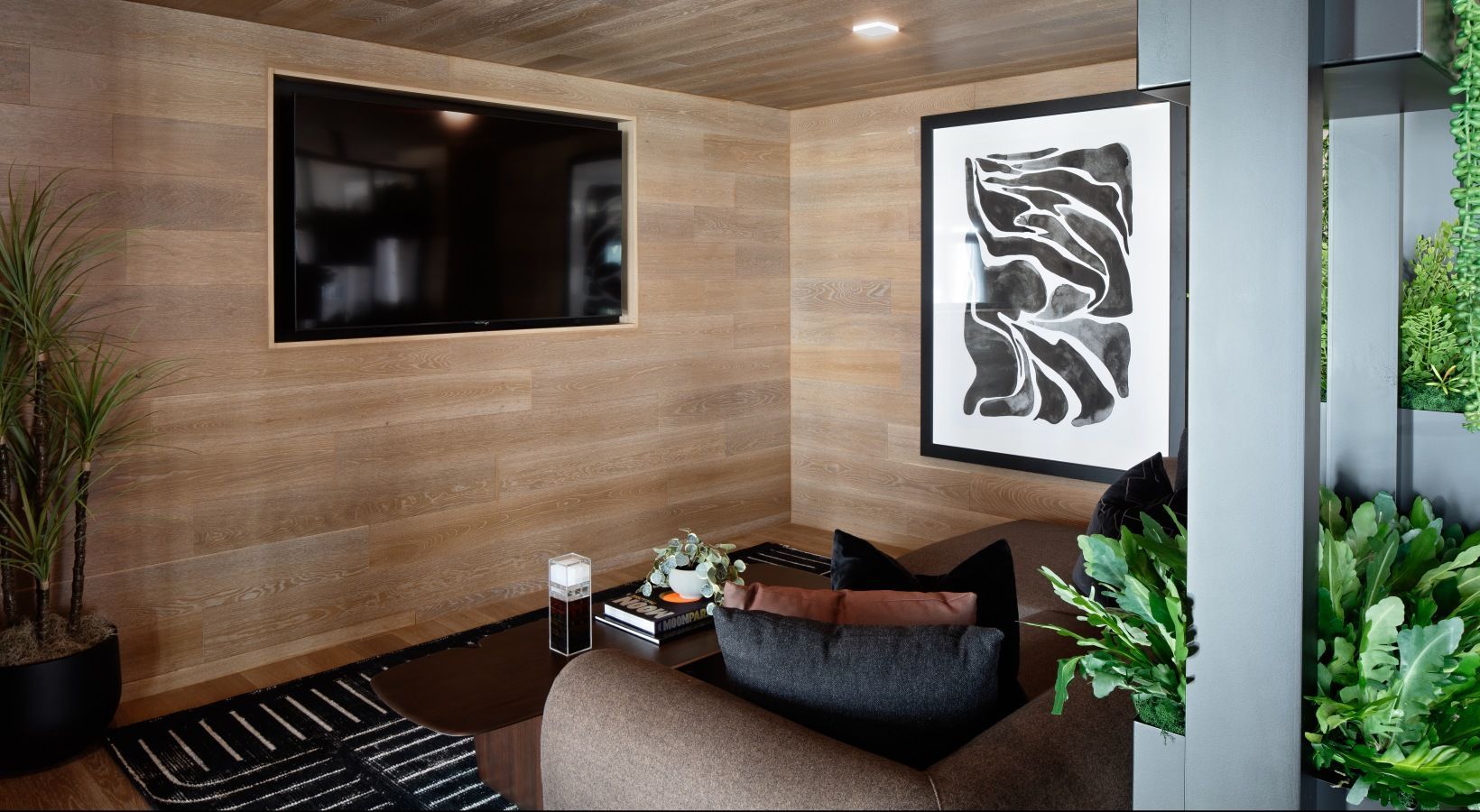 Resident lounge in a modern apartment community with a mounted TV, abstract art, wood panel walls, and plants at Sofia, offers apartments in Santa Clara, CA.