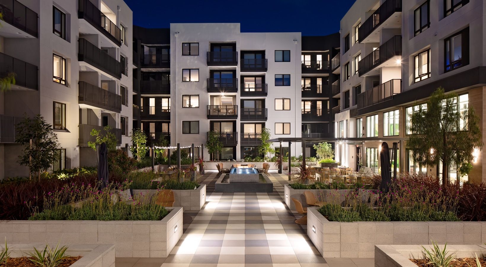 Night view of a modern apartment courtyard with a central fountain, planters, and outdoor seating at Sofia, offers apartments for rent in Santa Clara, CA.