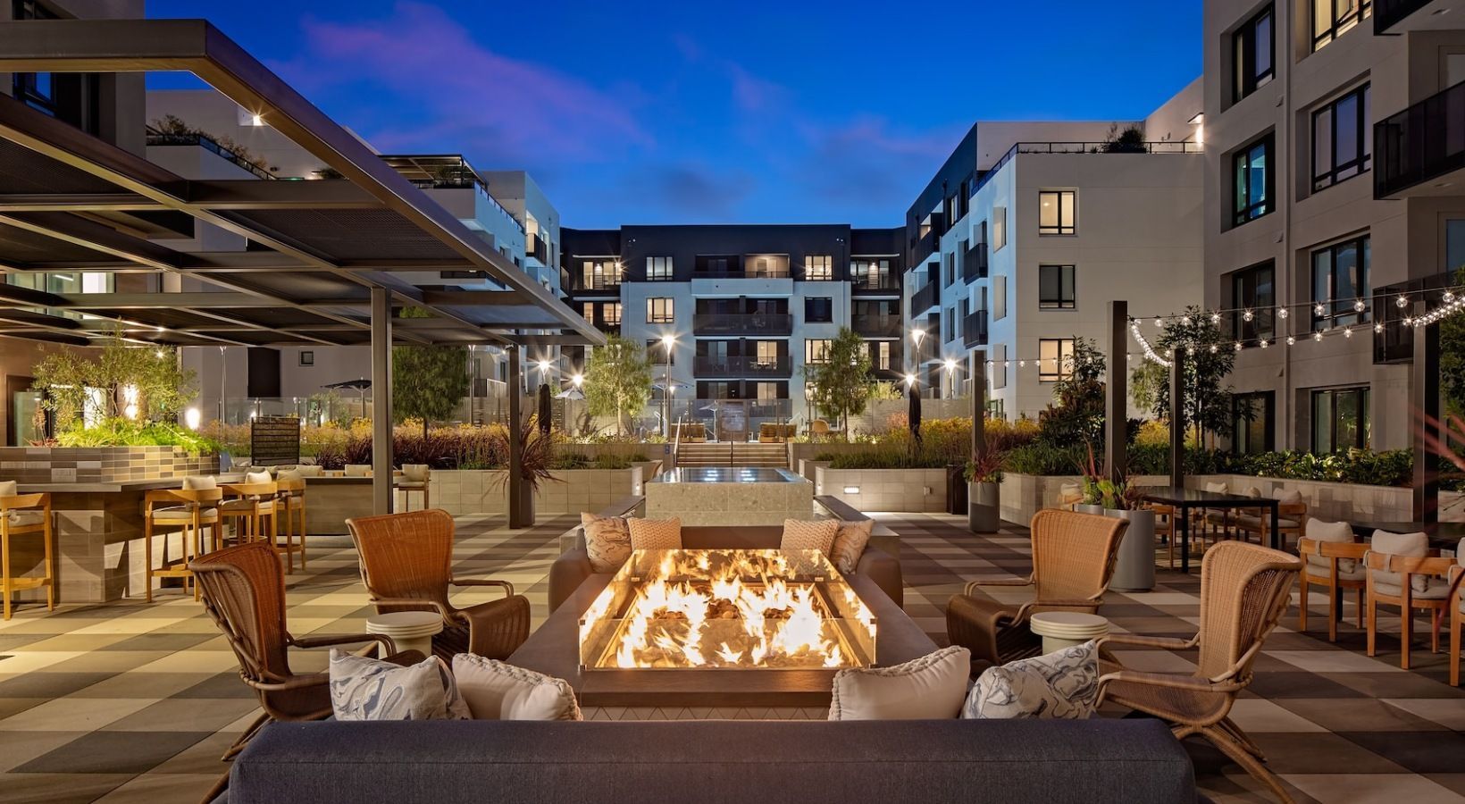 Courtyard at dusk with fire pit, seating, string lights, and modern apartment buildings.