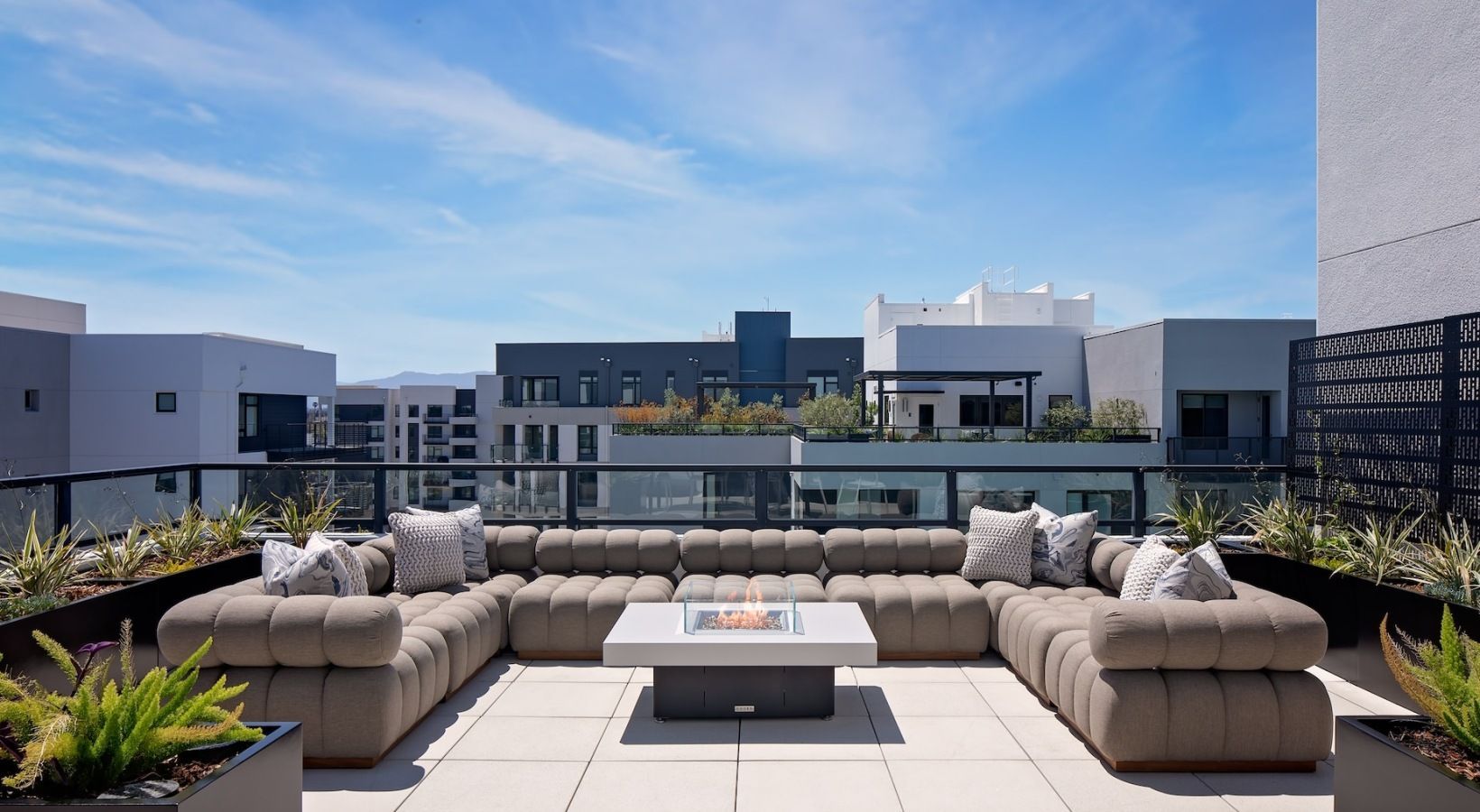 Rooftop patio with large sectional sofa, fire pit, and cityscape under a bright blue sky.