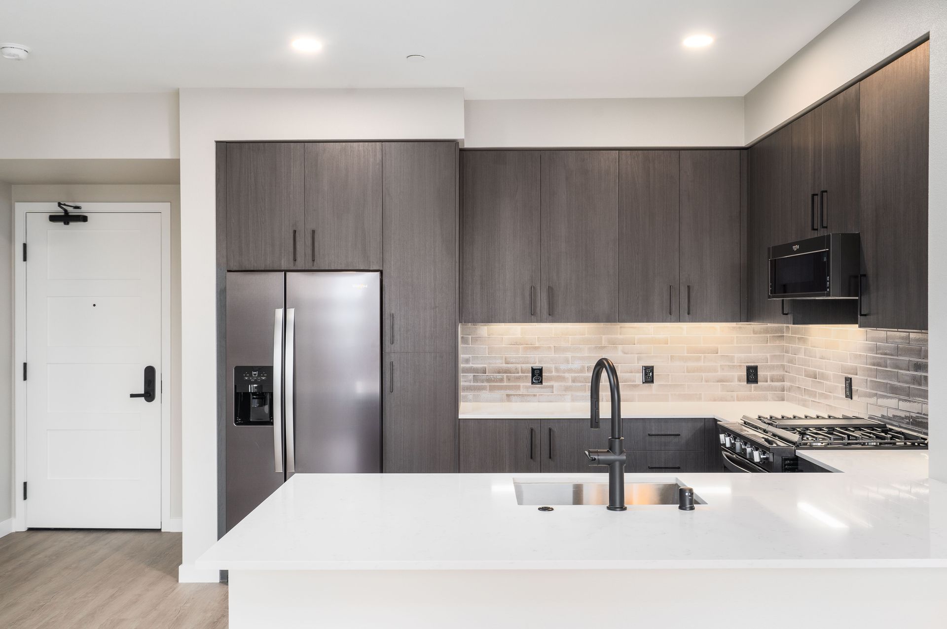 Modern kitchen with dark wood cabinets, stainless steel appliances, white countertop island, and white door at Sofia, offers studio, 1, and 2 bedroom apartments in Santa Clara, CA.