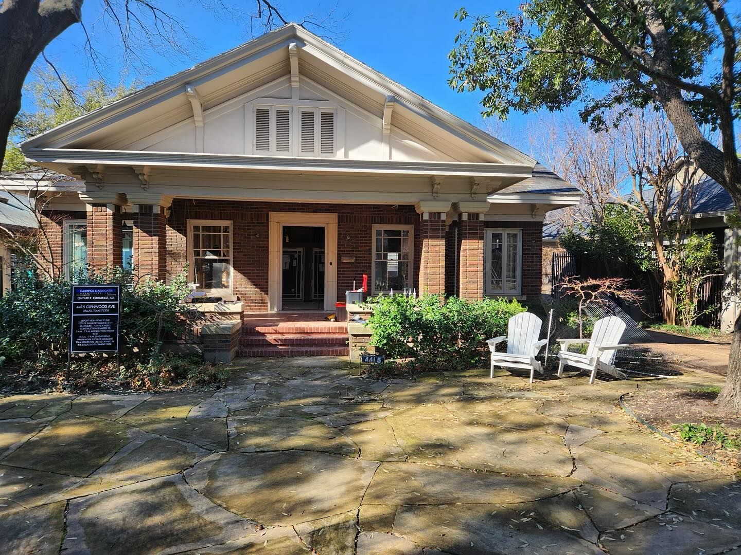 A brick house with white chairs in front of it on a sunny day.