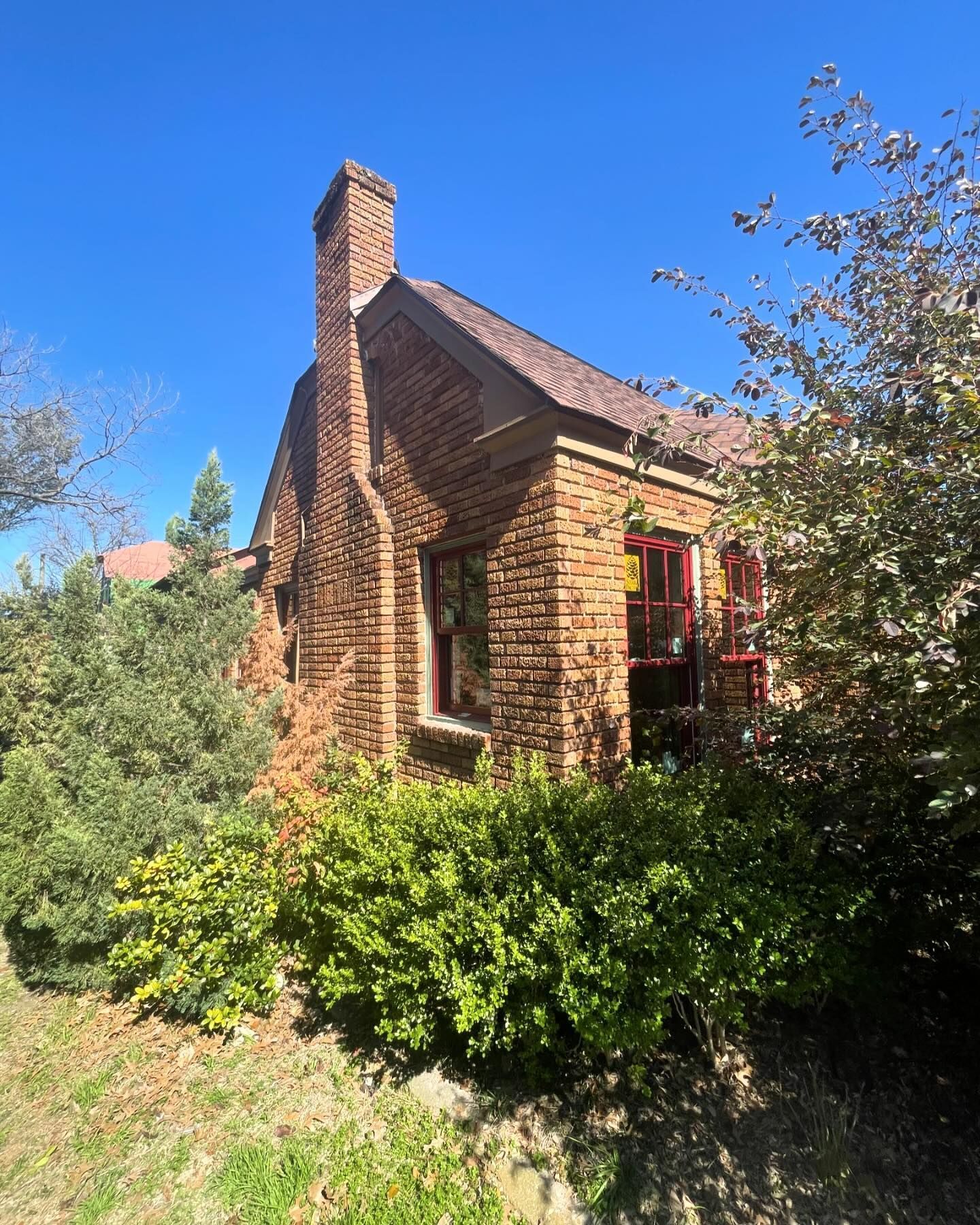 A small brick house with a chimney is surrounded by bushes and trees on a sunny day.