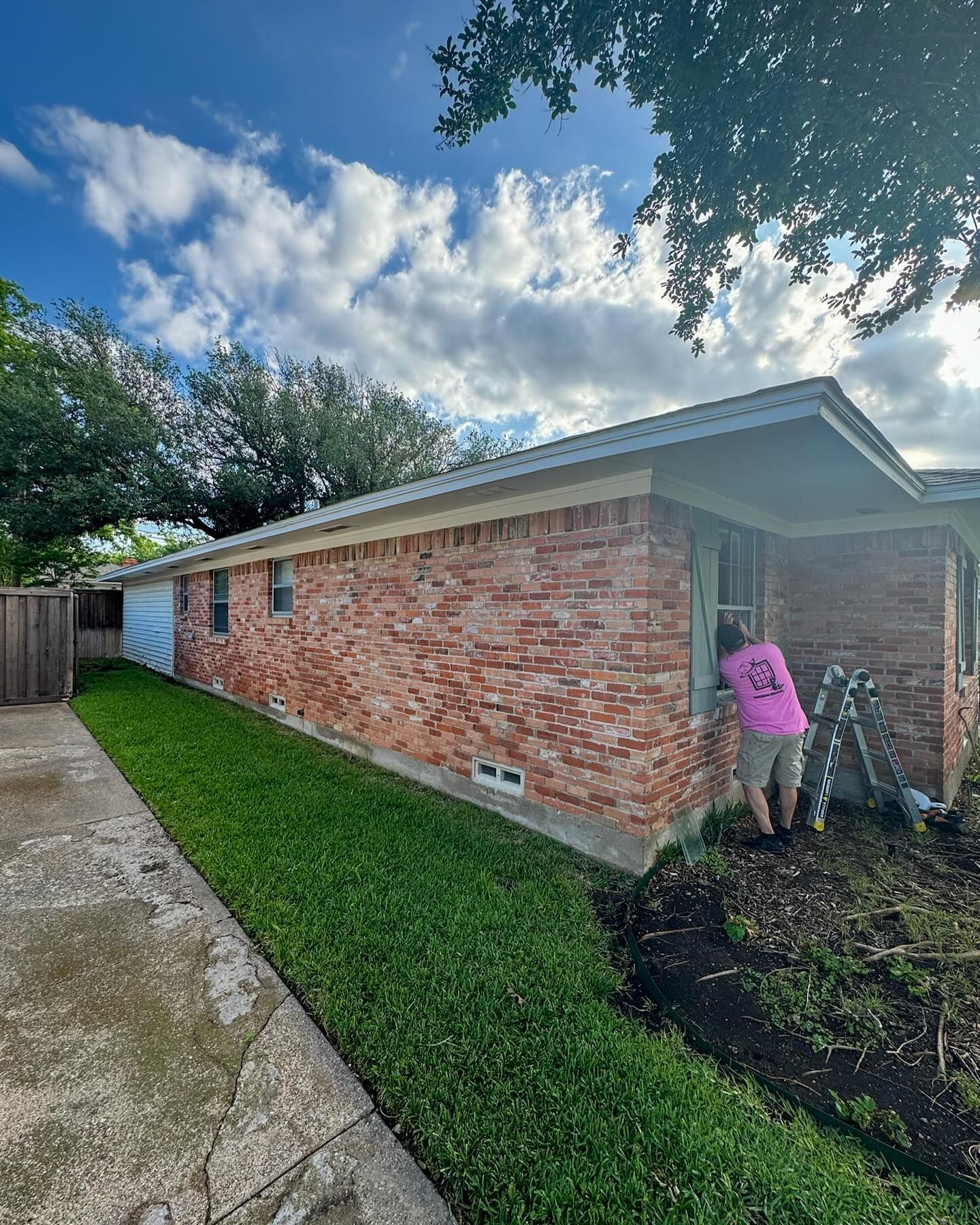 A woman in a pink shirt is standing in front of a brick house.