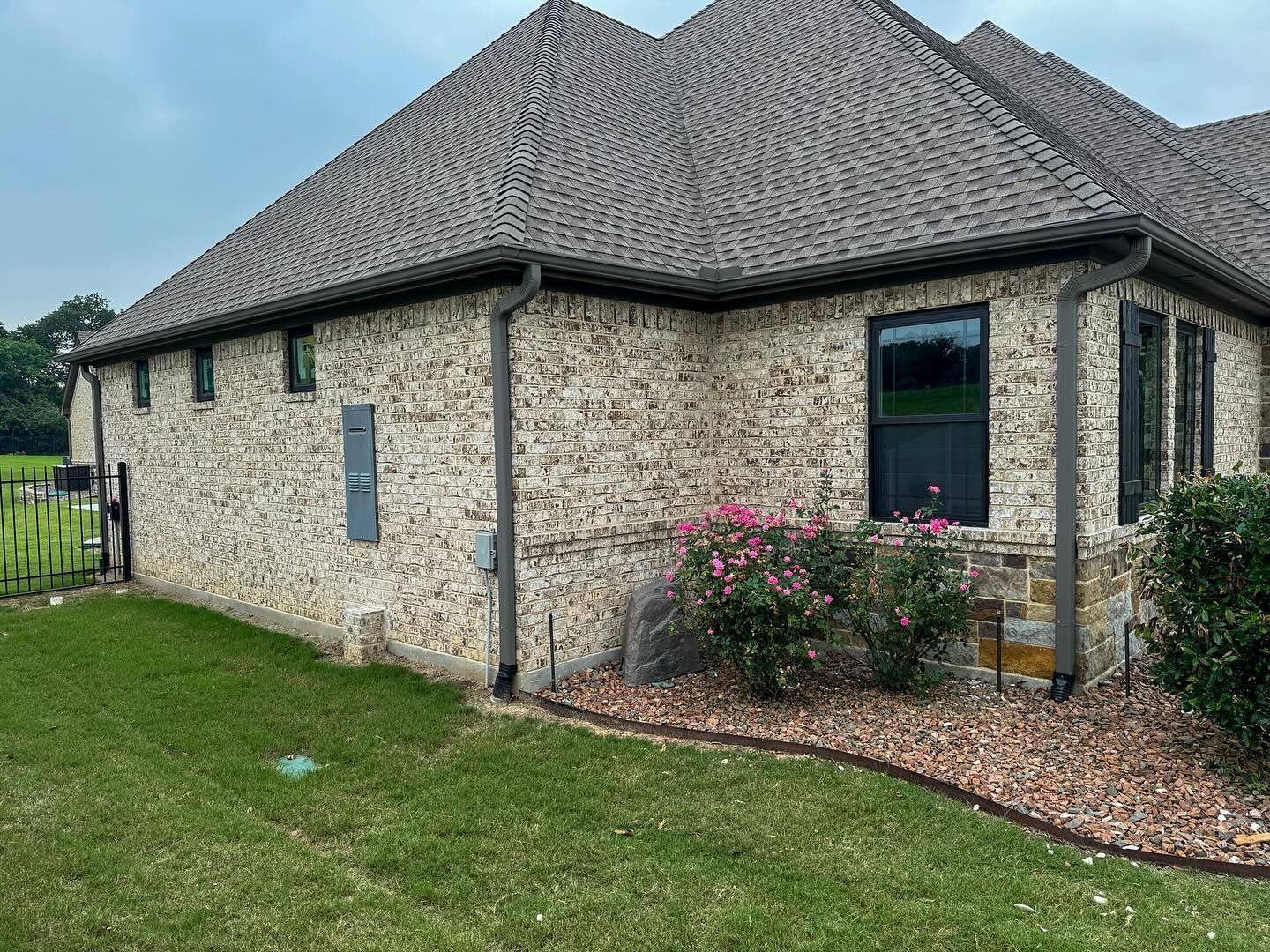 A large brick house with a gray roof and a lot of windows is sitting on top of a lush green lawn.
