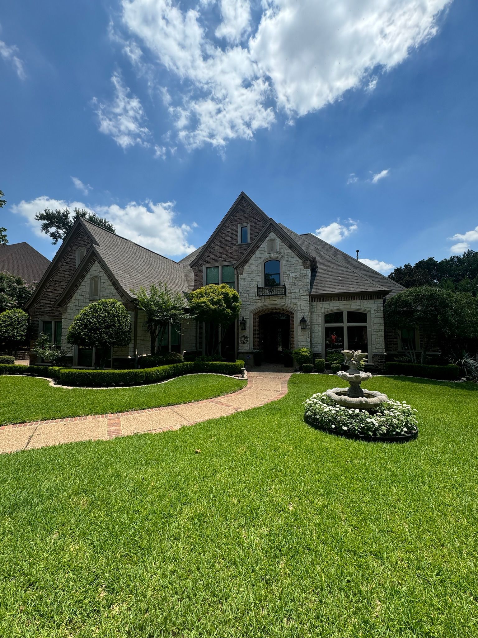 Stone house with steep gray roof, manicured lawn, and fountain under a bright blue sky.