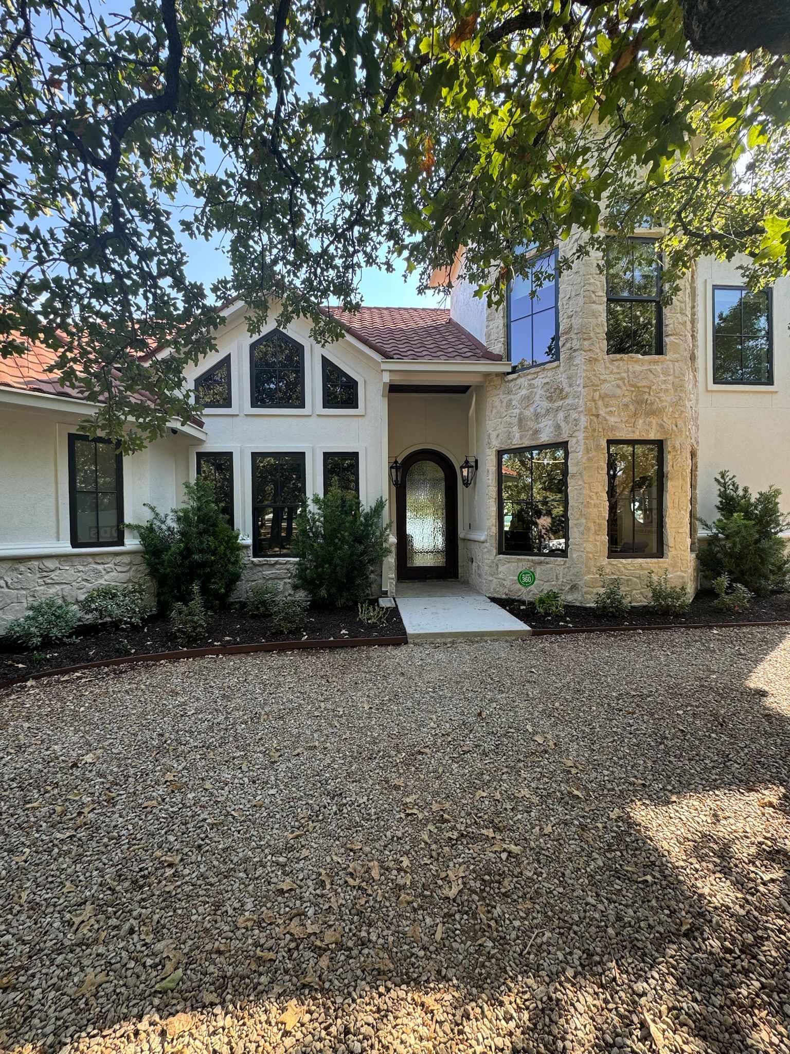Front view of a stucco house with a stone tower, arched entry, and gravel driveway under trees