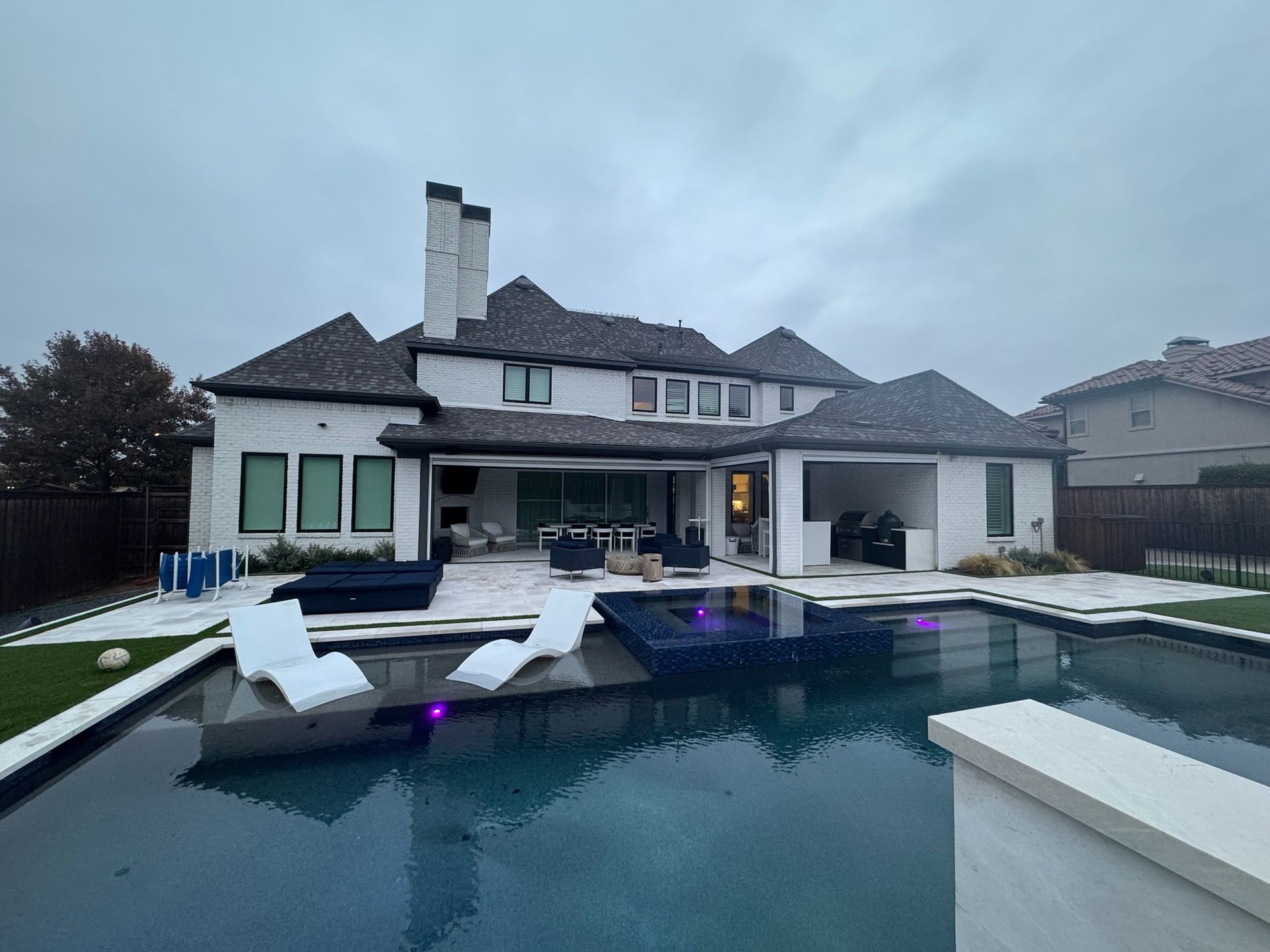 Backyard infinity pool with lounge chairs beside a large modern house at dusk