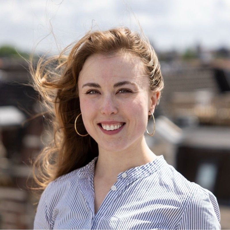 Woman with light brown hair smiles, wearing hoop earrings and a blue and white striped shirt outdoors.