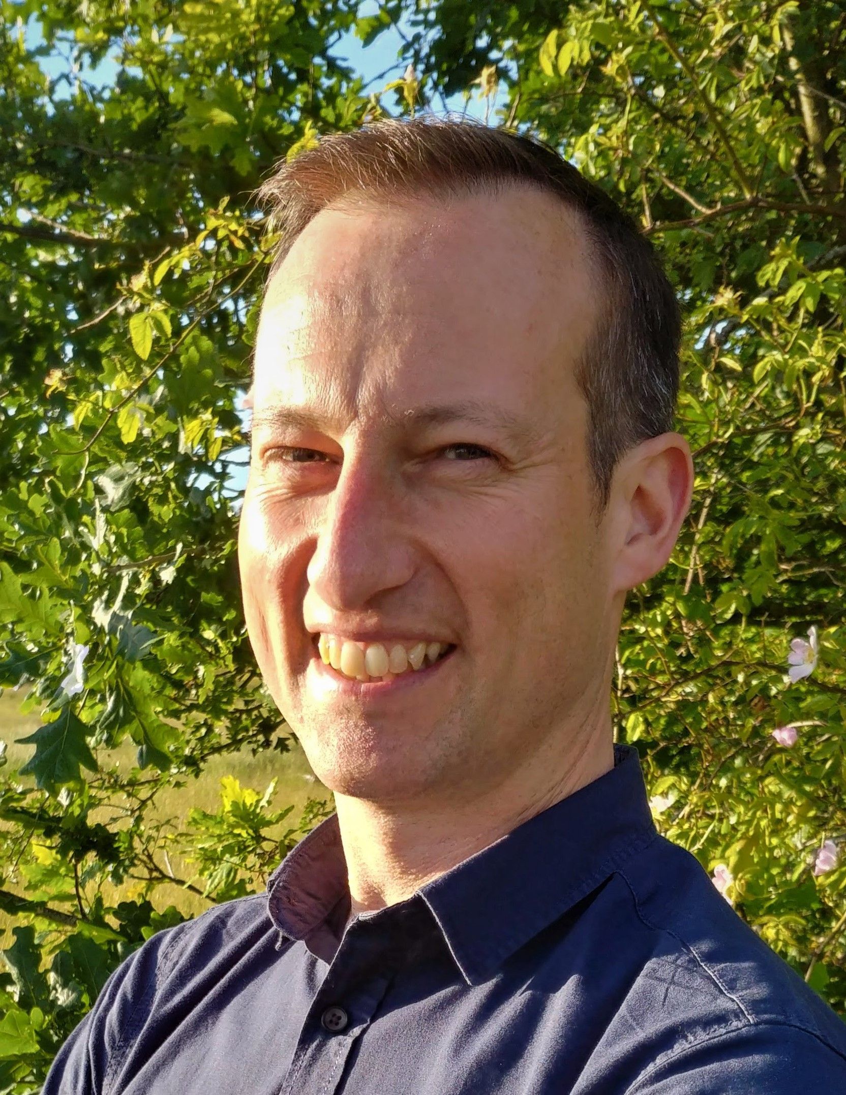A man in a blue shirt is smiling in front of a tree.