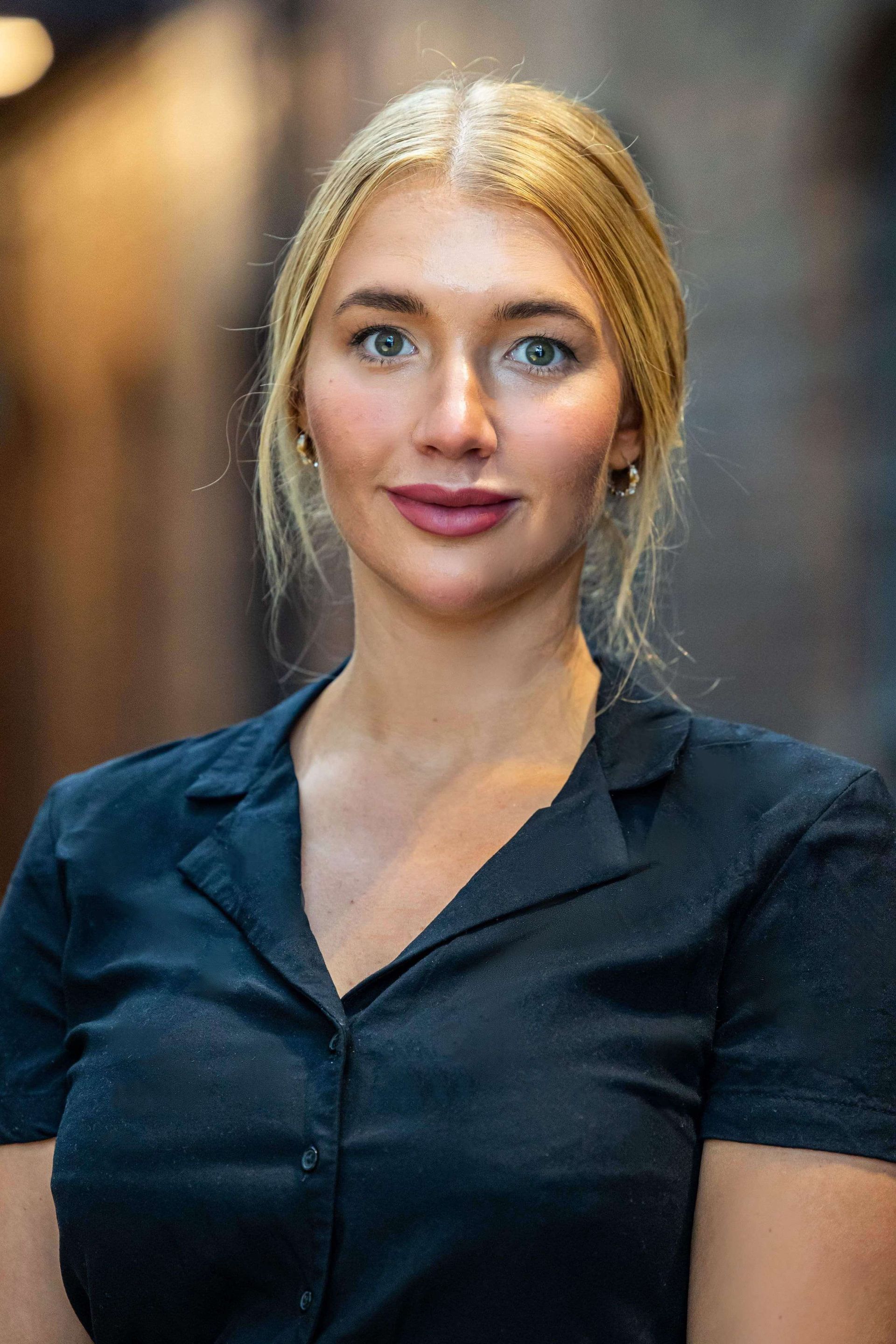 Blonde haired woman in black shirt, looking at camera, background blurred.