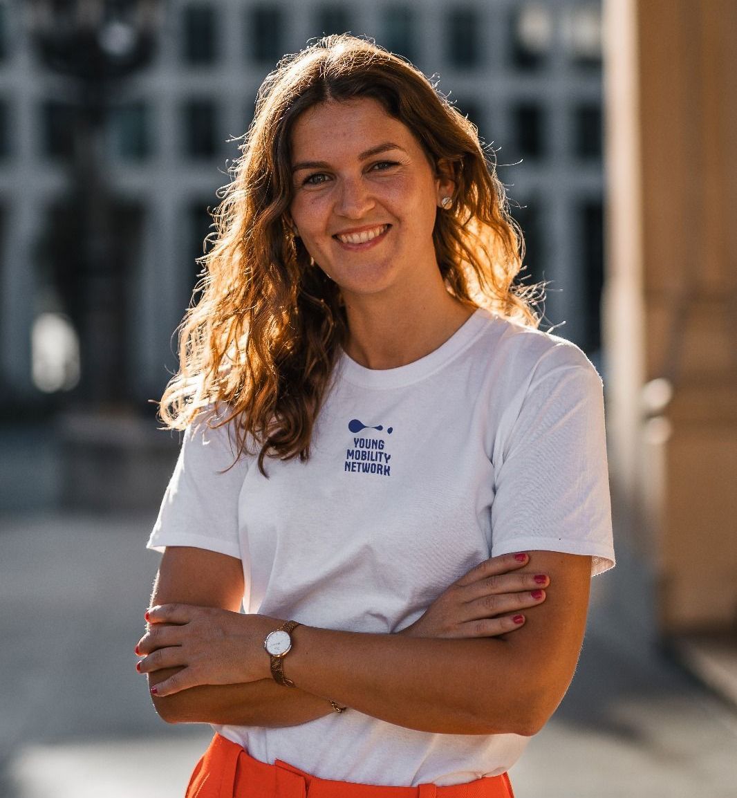 A woman wearing a white t-shirt and orange trousers is standing with her arms crossed and smiling.