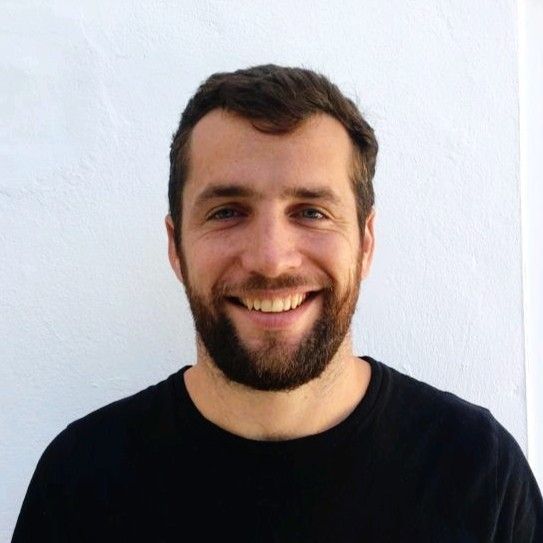 Man with a beard smiling in front of a white wall, wearing a black t-shirt.