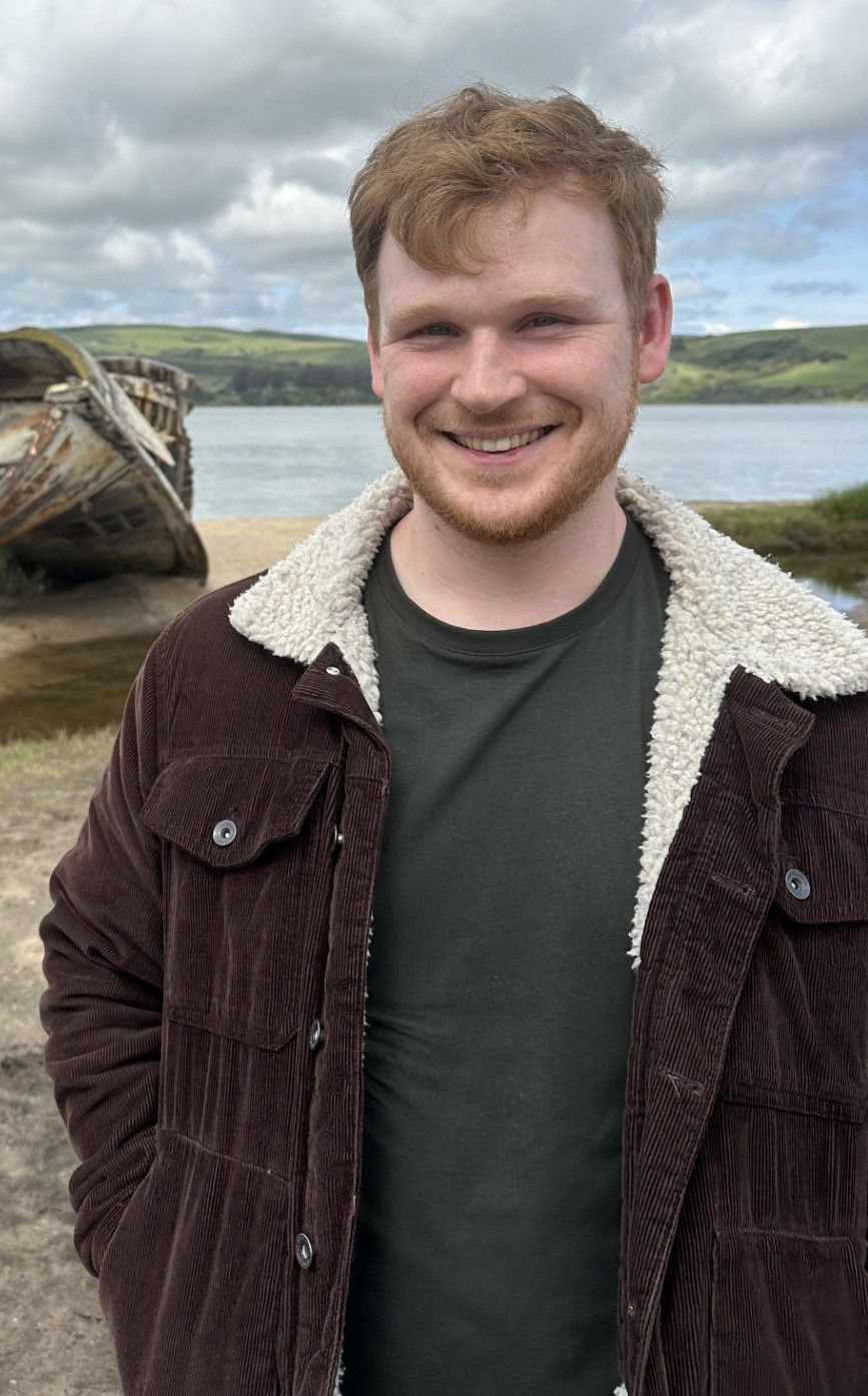 Man with red hair, smiling, wearing a brown corduroy jacket with a white collar, standing outside by water.