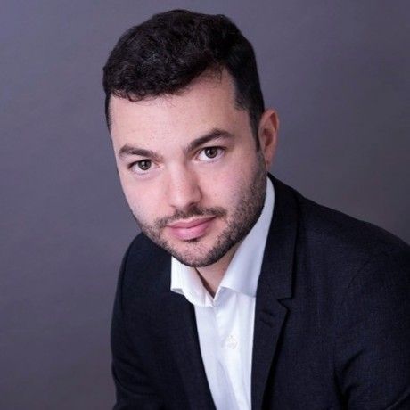 Man in black suit and white shirt, looking at the camera with a slight smile, against a gray background.