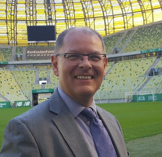 Man in suit smiles. Stands in front of green field and yellow seating in a stadium.