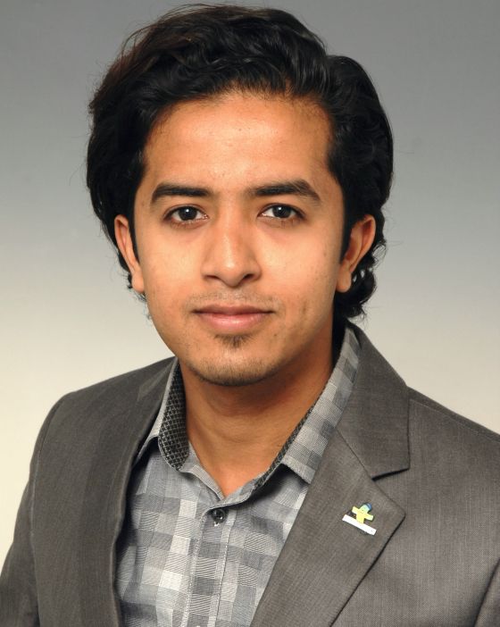 Man in a gray suit, wearing a checkered shirt, looking at the camera.