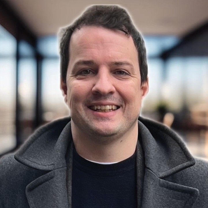 Smiling man wearing a gray coat and black sweater, indoors.