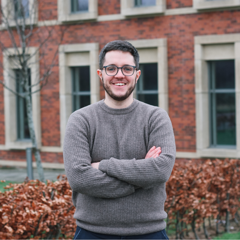 Man with glasses and a beard smiling with arms crossed in front of a brick building.