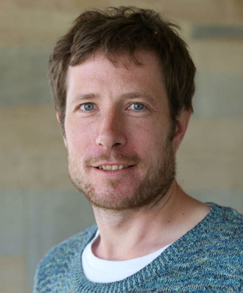 Man with blue eyes smiles slightly, wearing a blue sweater over a white t-shirt.