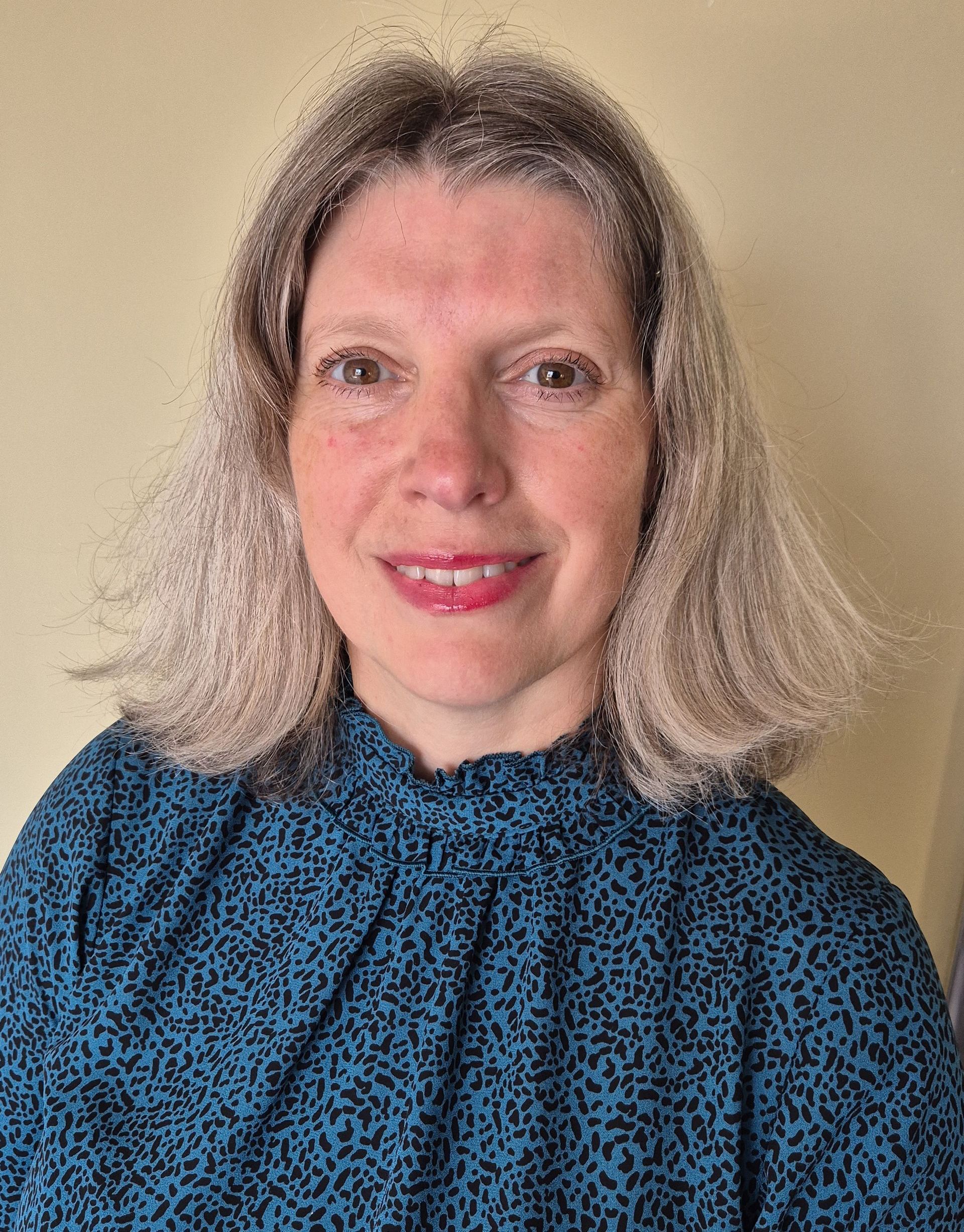 Woman with wavy blonde hair smiles, wearing a blue patterned shirt against a light yellow wall.